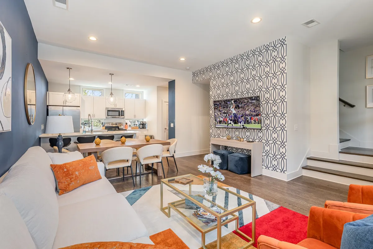 A vibrant living room featuring a plush sofa with colorful cushions, an adjacent dining area with a large table, decorative wallpaper, and a television screen. The space feels lively and designed for both entertainment and relaxation, highlighting the entertainment and dining aspects.