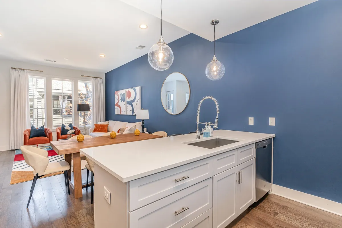 A wide-angle view of the open-plan living, dining, and kitchen area. The scene shows a kitchen island with bar stools, a contemporary dining table, and a comfortable seating area with modern wall art. Natural light fills the space, emphasizing its welcoming and sociable atmosphere.