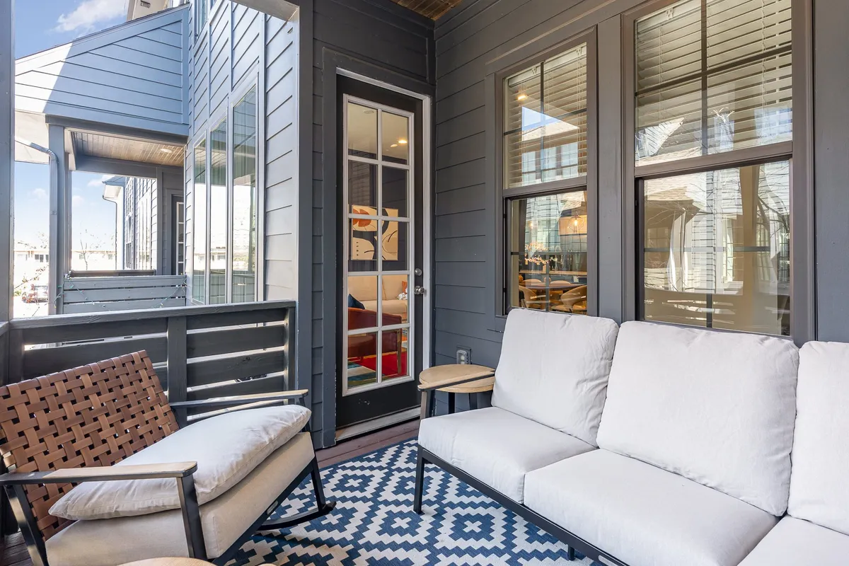 This image depicts a balcony or patio space with comfortable outdoor seating, a decorative rug, and a door leading inside. The setup suggests a welcoming and relaxed atmosphere ideal for lounging, socializing, or unwinding. The greenery and open air evoke a sense of tranquility and connection to the surrounding neighborhood.
