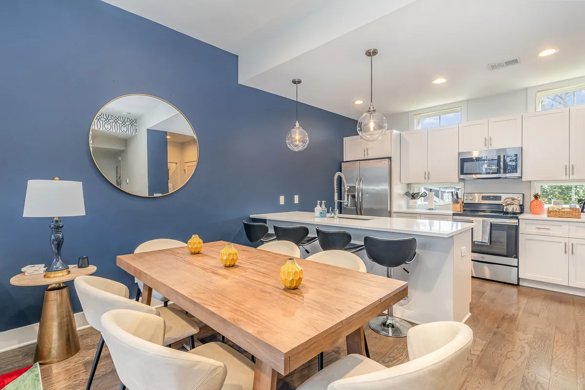 A well-lit dining area with a dining table and kitchen island, round mirror, and stylish pendant lights. The space is arranged for gatherings, meals, and casual get-togethers, exuding an inviting, communal vibe.