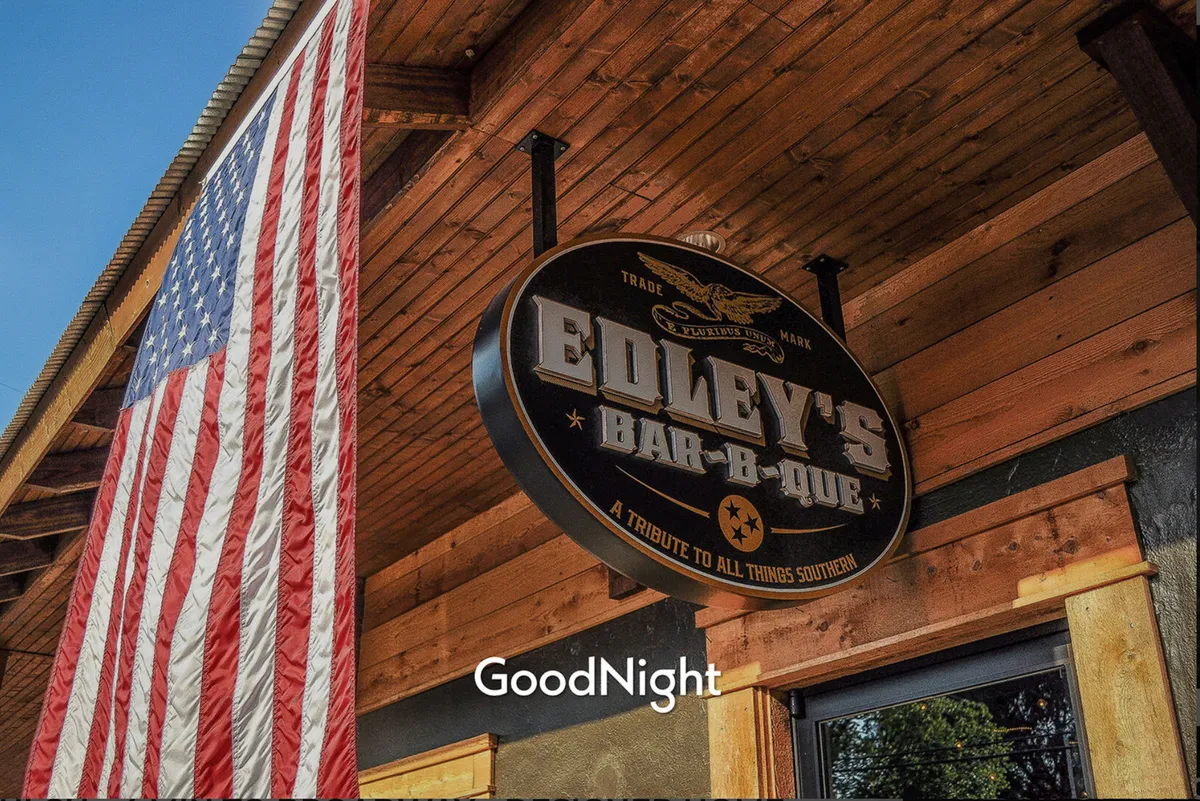Exterior view of Edley’s Bar-B-Que, showing the iconic sign and an American flag above the entrance. The restaurant exterior radiates a friendly, inviting atmosphere, suggesting a popular local dining spot for families and groups.