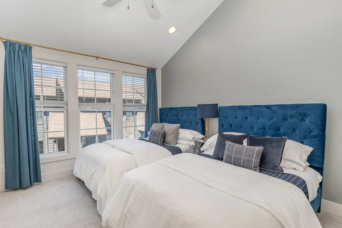 A spacious bedroom with two beds featuring blue tufted headboards, a shared bedside lamp, and large windows. The setting exudes calm and comfort, making it perfect for guests who want both togetherness and personal space.