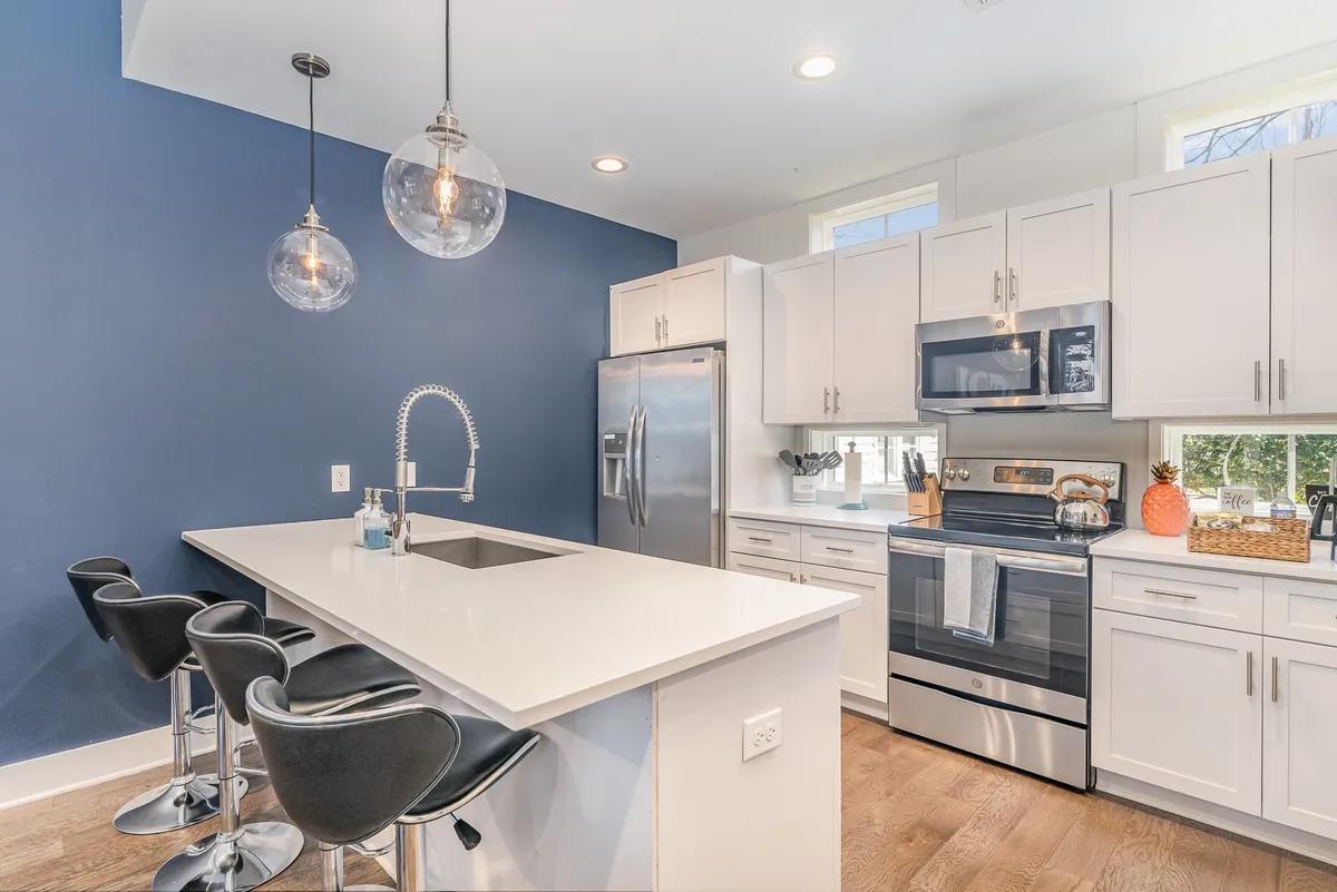 A contemporary kitchen highlighted by a central island with bar stools, pendant lights, stainless steel appliances, and a blue accent wall. The setup is ideal for group cooking, casual snacks, and mingling, with a welcoming, modern feel.