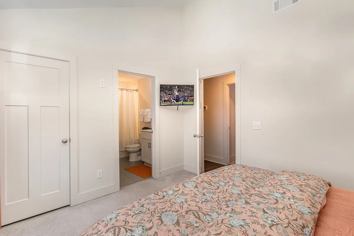 A well-appointed bedroom with floral bedding on the bed, a wall-mounted television, and doorways leading to an en-suite bathroom and hallway. The room feels inviting and well-equipped for modern comforts.