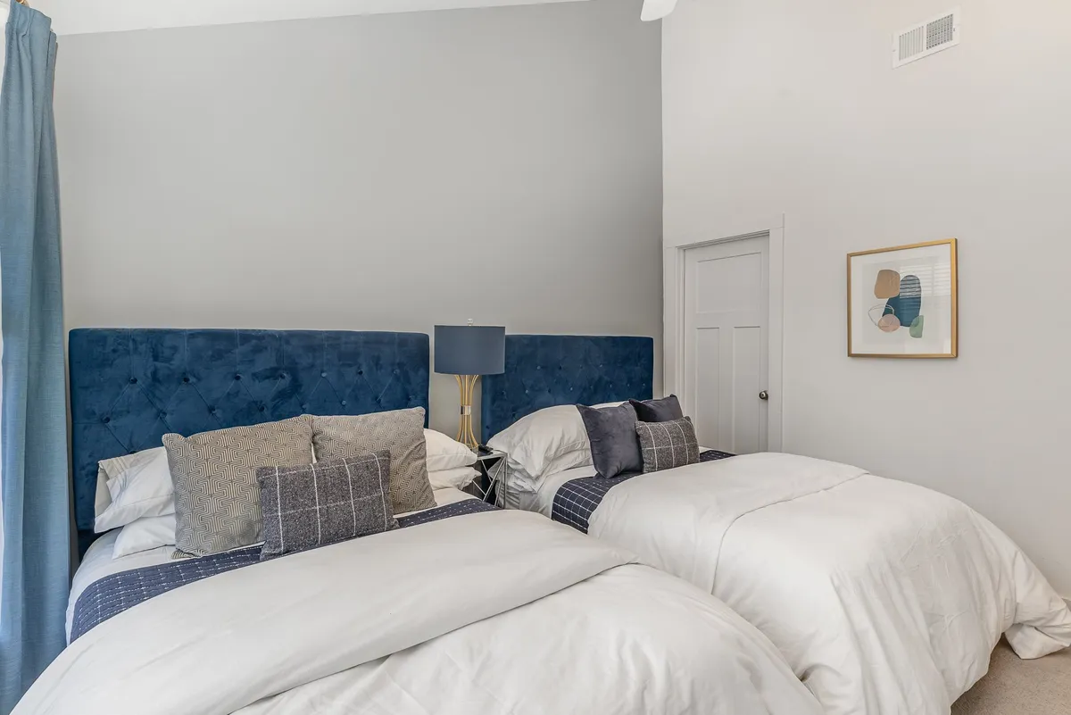 The bedroom features cozy blue velvet headboards, crisp white bedding, decorative pillows, a nightstand with a lamp, and artwork on the wall. The relaxing atmosphere is perfect for restful nights and quiet moments.