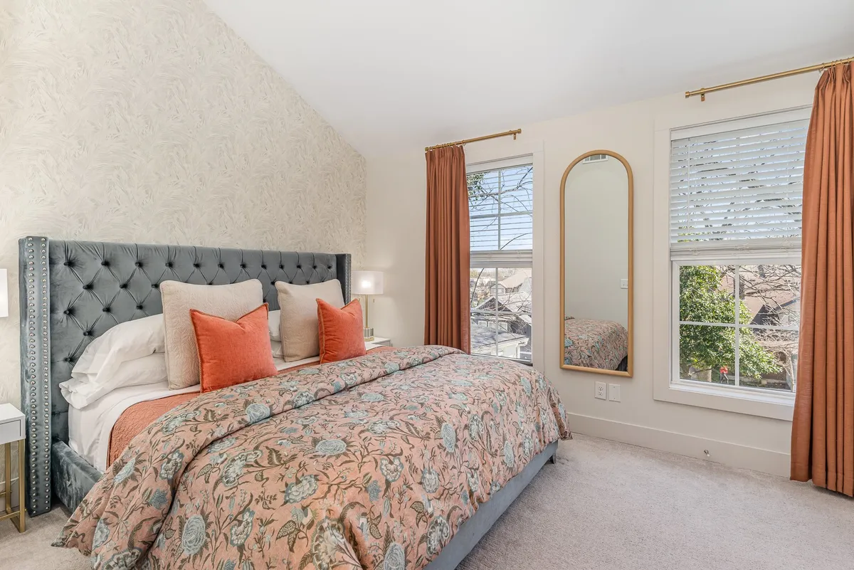 A cozy bedroom scene featuring a bed dressed with decorative pillows, a large window with soft curtains letting in natural light, and a full-length mirror. The neutral tones and serene styling evoke a sense of tranquility and comfort.