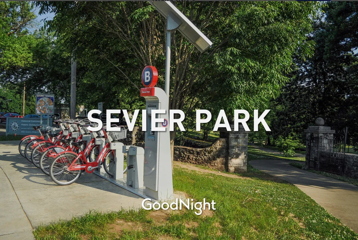 This exterior image features a bike rental station at the entrance to a lush, green park. Park signage is visible, and the surrounding area is filled with leafy trees and well-maintained paths, creating an inviting space for outdoor activities such as biking, walking, and picnicking.